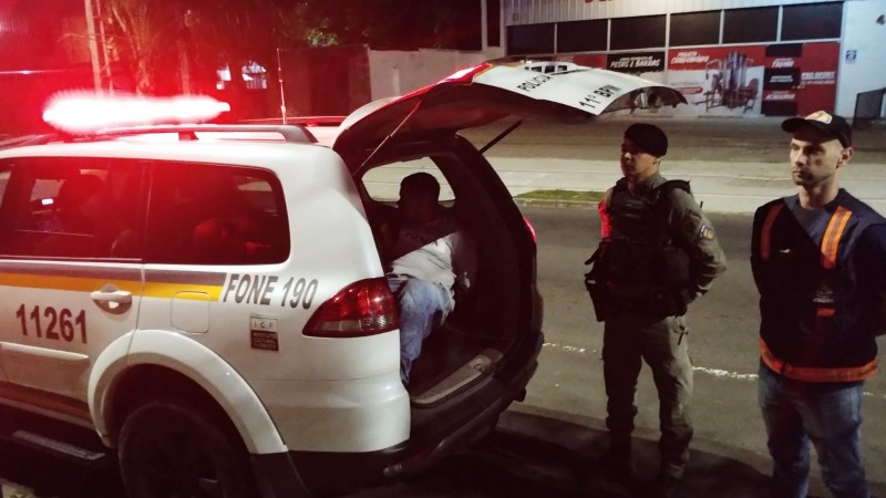 Foto horizontal colorida mostra um agente de trânsito e um policial militar parados ao lado de um carro da polícia, com um preso na parte de trás do veículo