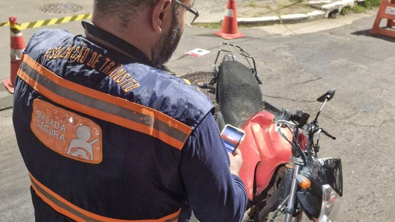 Foto horizontal colorida mostra agente de transito com colete da Balada Segura de costas olhando para o celular, de frente para uma moto vermelha. Se vêem cones e cavaletes ao fundo