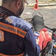 Foto horizontal colorida mostra agente de transito com colete da Balada Segura de costas olhando para o celular, de frente para uma moto vermelha. Se vêem cones e cavaletes ao fundo