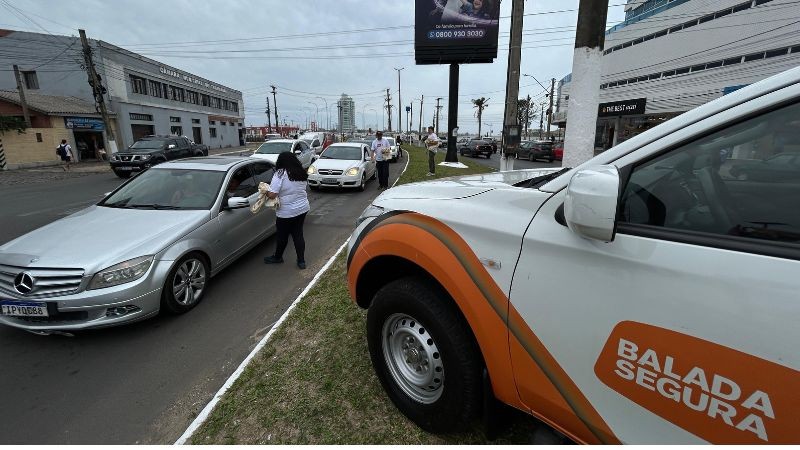 Foto horizontal colorida mostra o cenário de uma avenida movimentada com a vitura da Balada Segura parada no canteiro e agentes conversando com motoristas pela janela dos carros