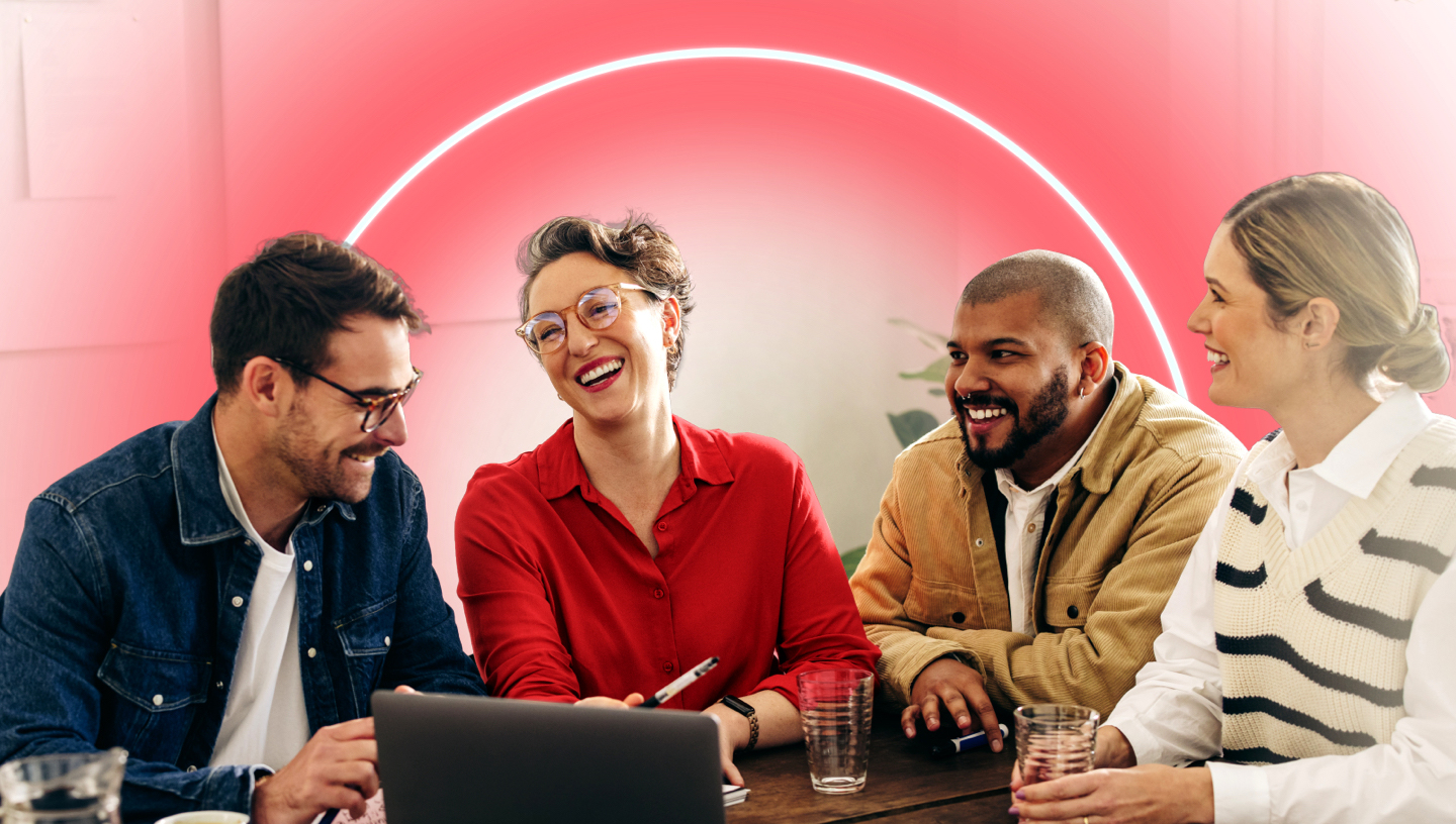 visual: A group of four professionals sit at a small table. They surround an open laptop as a few smile and others laugh cordially.