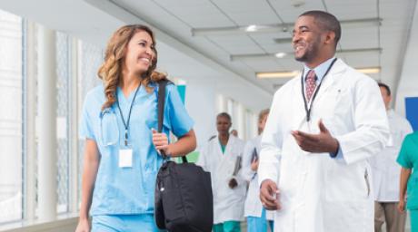 Doctor in white coat and person in scrubs talking while walking down hospital hallway