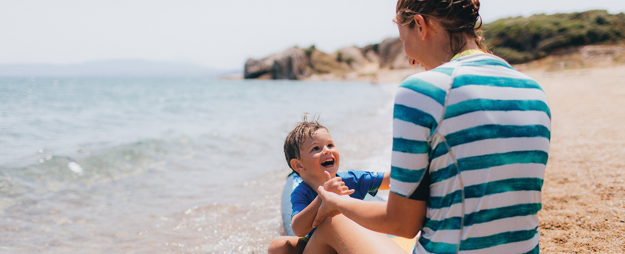 A mom and her young son sit on a surfboard in the sand. 