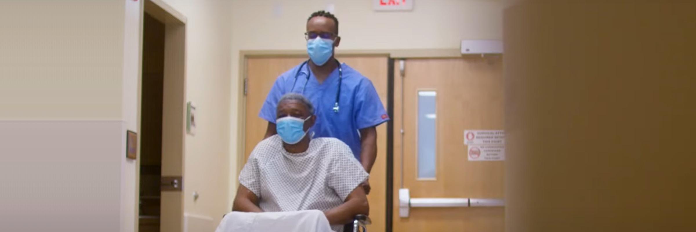 Black male nurse with glasses and a mask wheels a older black man in a wheelchair, gown, and mask down a hospital corridor.