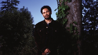 Photo of Kaytranada wearing a black shirt against a blue sky and desert background