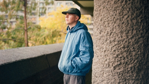 Nectax wearing a light blue rain coat and cap, standing on a large concrete balcony looking out at some trees