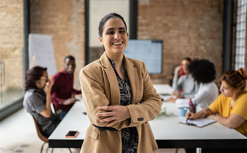 Une femme dans un blazer beige sourit les bras croisés dans un bureau, avec des collègues qui se rencontrent à une table derrière elle.