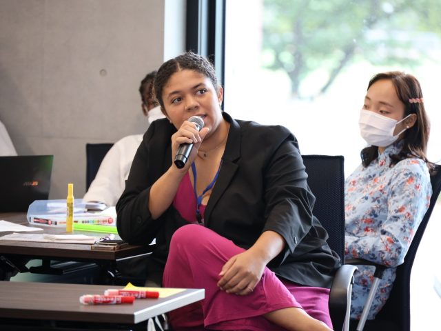 Chante Hardaway wearing a pink jumpsuit and blazer speaking into a microphone