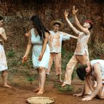 Foto dos atores do grupo de teatro. Duas mulheres e três homens. Vestidos de branco, sujos de poeira marrom. O local da foto é um terreno com terra, algumas poucas plantas rasteiras e um paredão de terra que toma todo o fundo da foto.