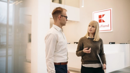 Eine Mitarbeiterin ist am Empfang mit ihrem Kollegen im Gespräch. Sie stehen vor einem weißen, modernen Tresen. Im Hintergrund ein großes Kaufland-Logo.