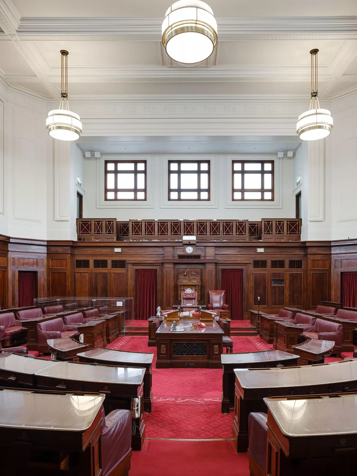 A view into the Senate Chamber looking straight into the room with benches in a U-shape on either side of the centre table. The carpet is a rich red and the seats are red leather. Art deco lights hang from the ceiling and wood timber panels surround the room. 