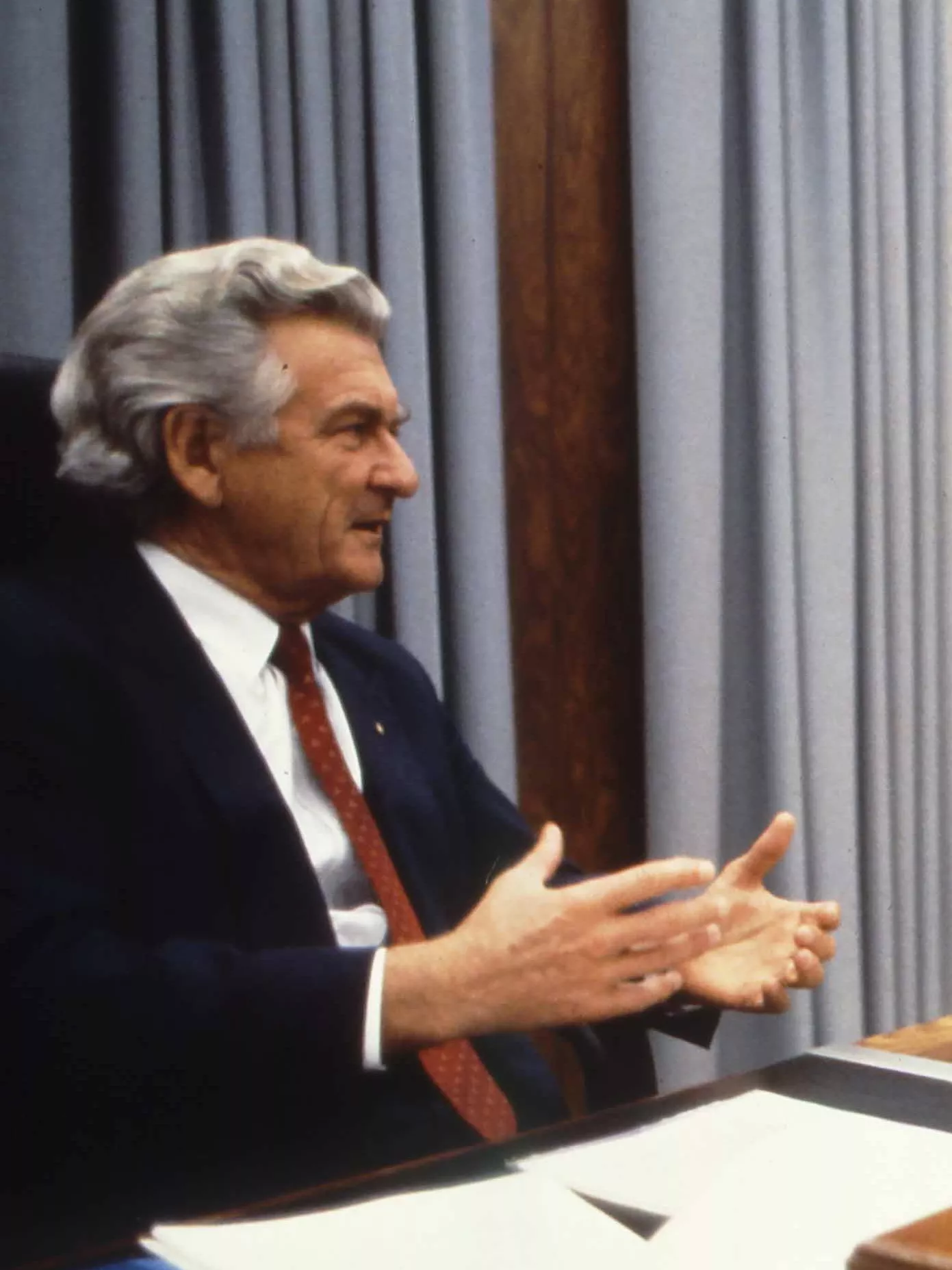 This colour photograph captures a conversation between Prime Minister Bob Hawke seated on the left and Treasurer Paul Keating seated on the right. The photo shows the corner of the desk with Hawke sitting behind the desk while Keating sits to the side with both men facing each other. Both are dressed in black suits with white shirts and red ties contrasting the pale blue curtains in the background.  