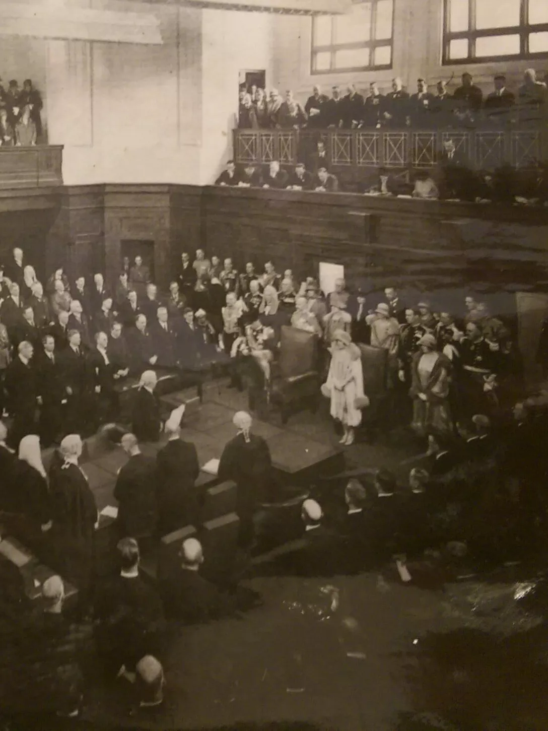 This black and white photograph shows the Duke of York reading a message from his father the King in the Senate Chamber. The room is crowded with at least 200 people, mostly men with some women all wearing their best. Men are dressed in suits, military uniform or gowns and wigs while the women wear formal dresses with hats and gloves. The crowd is in a horseshoe shape around the central table while the Duke stands at the head of the table.