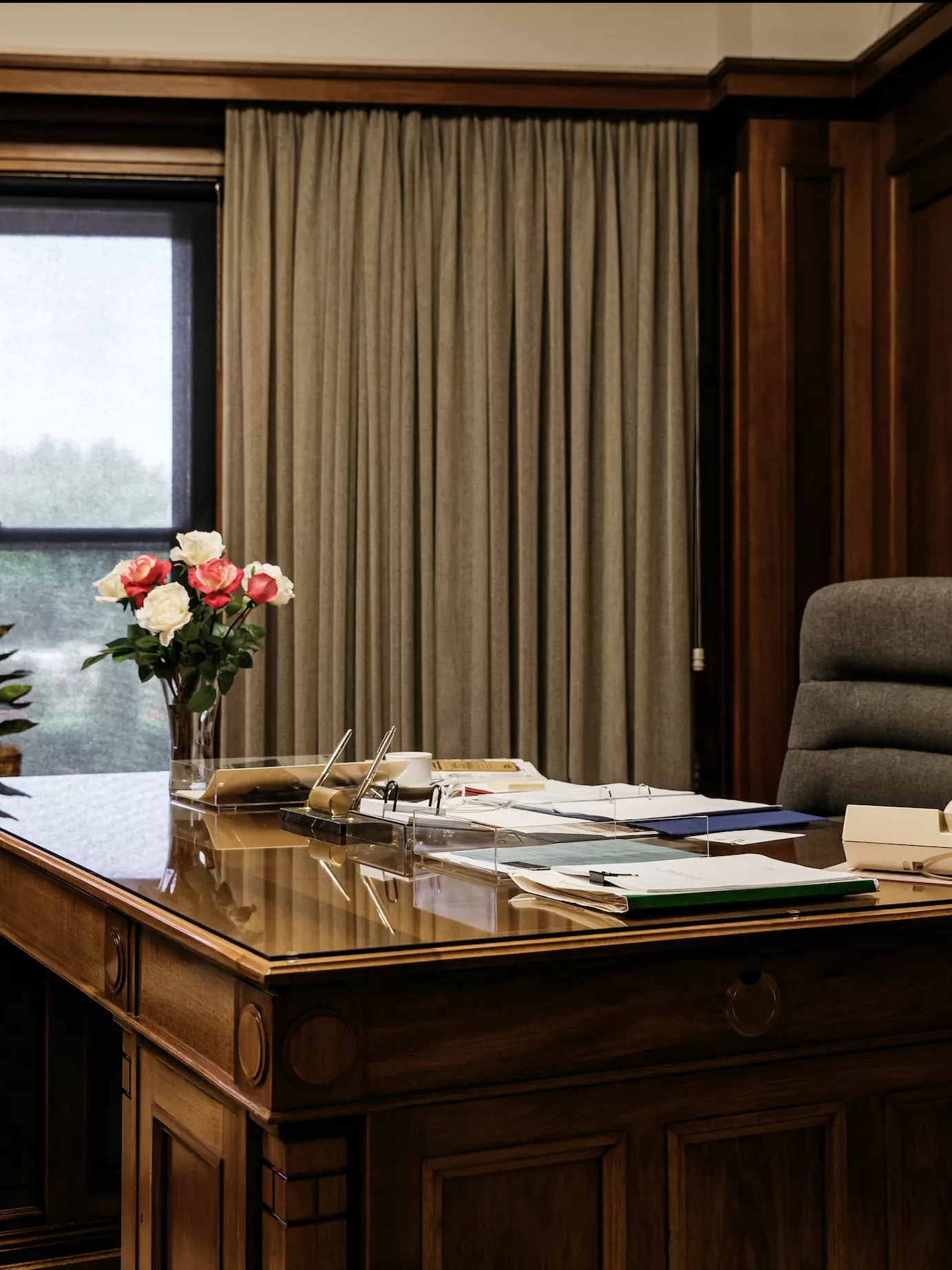 A large timber desk covered in folders and notes, with a vase with roses on it. 