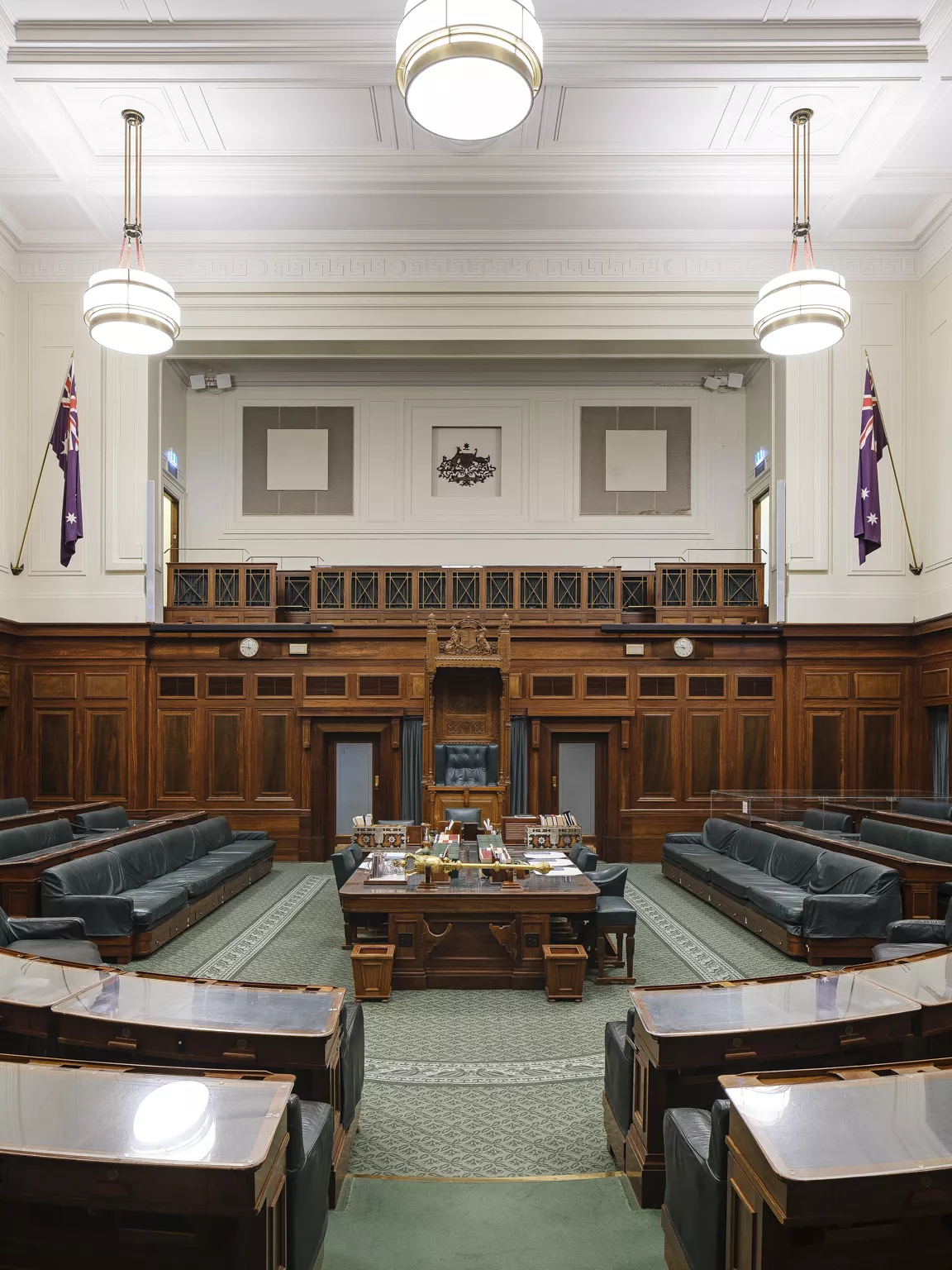 A green carpeted room with rows of wooden benches in a U shape facing a central table and the large, ornate Speaker's Chair. 