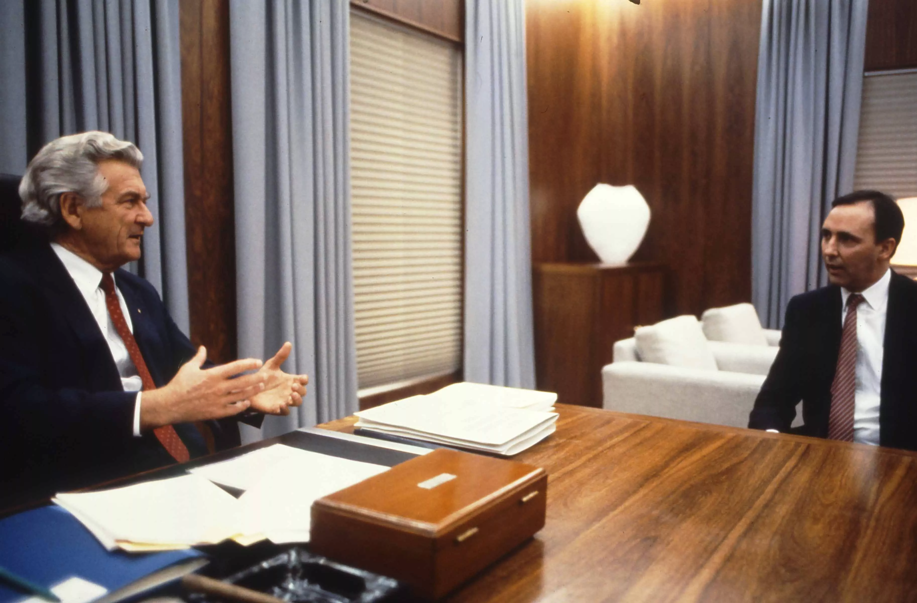 This colour photograph captures a conversation between Prime Minister Bob Hawke seated on the left and Treasurer Paul Keating seated on the right. The photo shows the corner of the desk with Hawke sitting behind the desk while Keating sits to the side with both men facing each other. Both are dressed in black suits with white shirts and red ties contrasting the pale blue curtains in the background.  