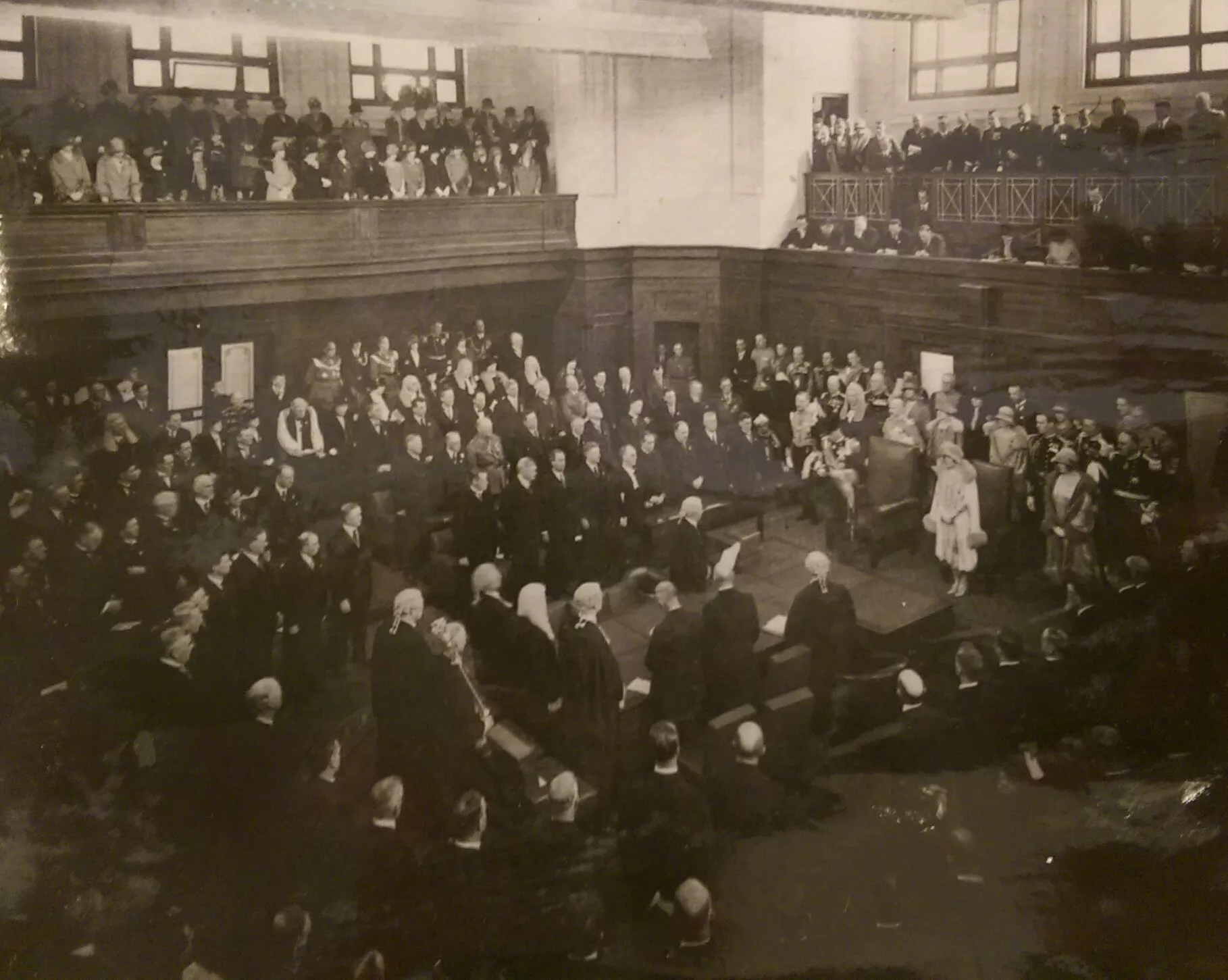 This black and white photograph shows the Duke of York reading a message from his father the King in the Senate Chamber. The room is crowded with at least 200 people, mostly men with some women all wearing their best. Men are dressed in suits, military uniform or gowns and wigs while the women wear formal dresses with hats and gloves. The crowd is in a horseshoe shape around the central table while the Duke stands at the head of the table.