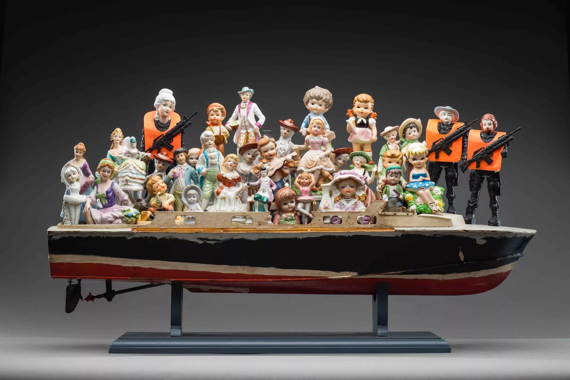 A porcelain statue of a boat filled with men, women and children in formal old fashioned outfits wearing bonnets, waist coats and bows and hats. There are 3 figurines win all black with orange vests and machine guns. 