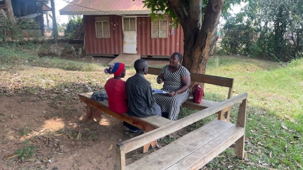 Three people talk on a bunch under a tree outside