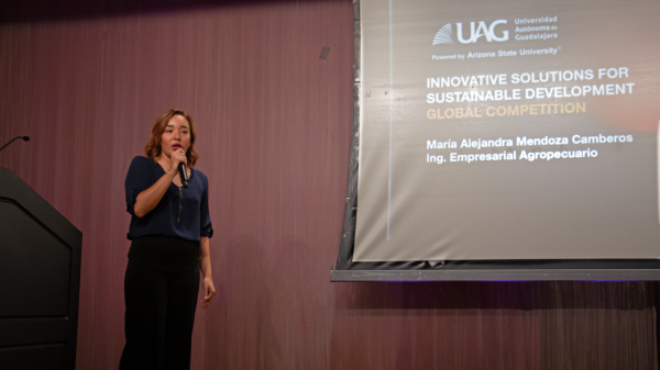 Woman speaking into a microphone in front of a screen with a presentation slide project onto it.