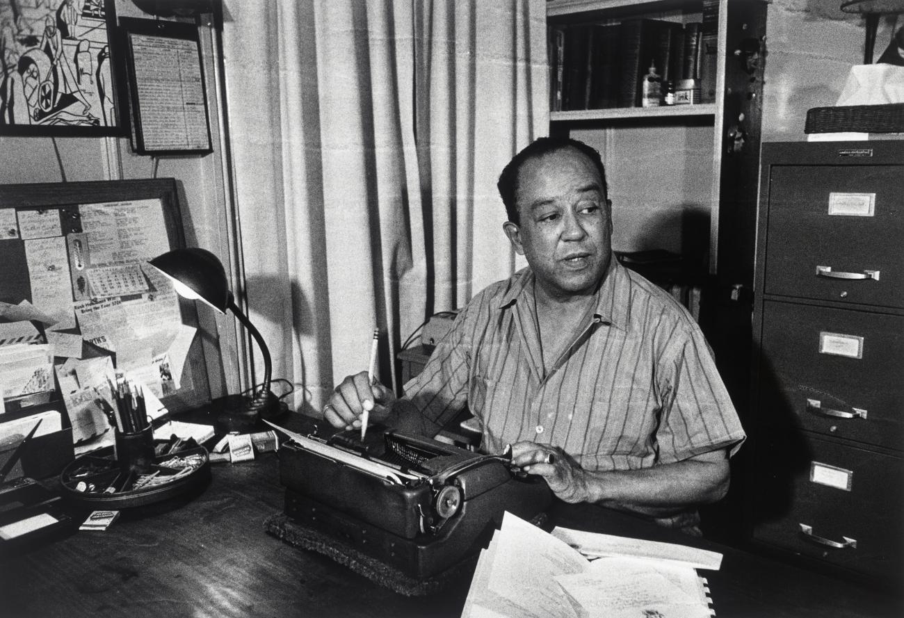 A photograph of Langston Hughes siting at a desk in front of a typewriter. 