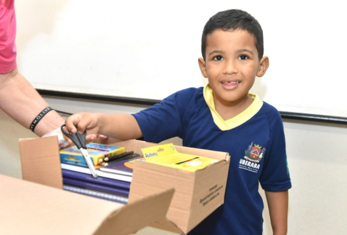 A imagem mostra uma criança com uma camisa azul e gola amarela segurando uma tesoura ao lado de uma caixa cheia de materiais escolares, com a ajuda de uma mão adulta.