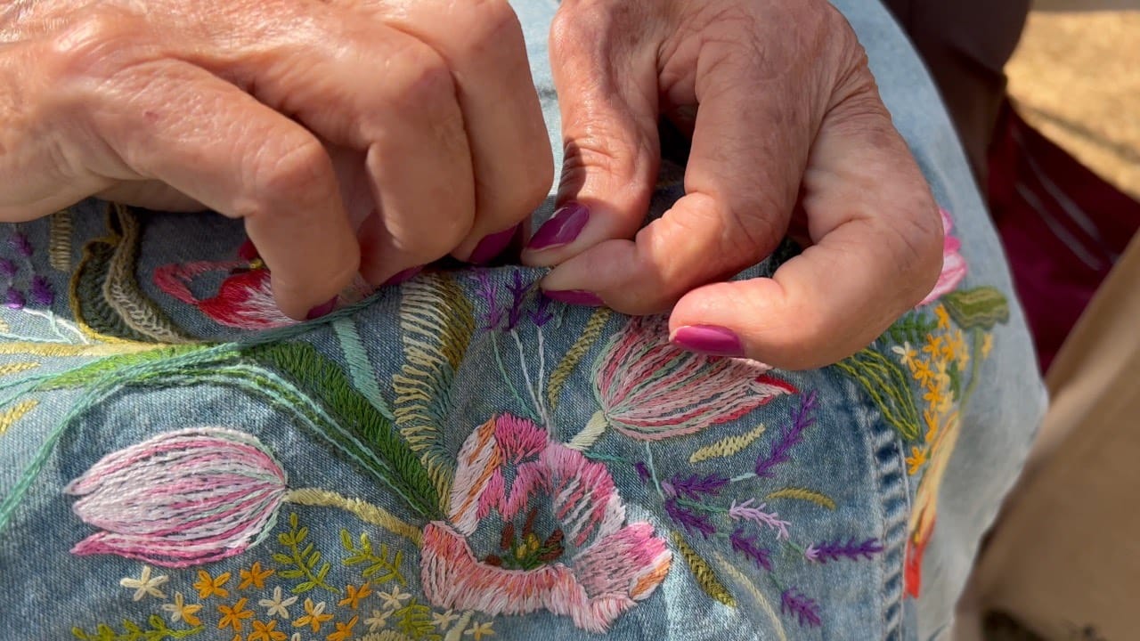 A imagem mostra duas mãos fazendo um bordado de rosas em um tecido azul 