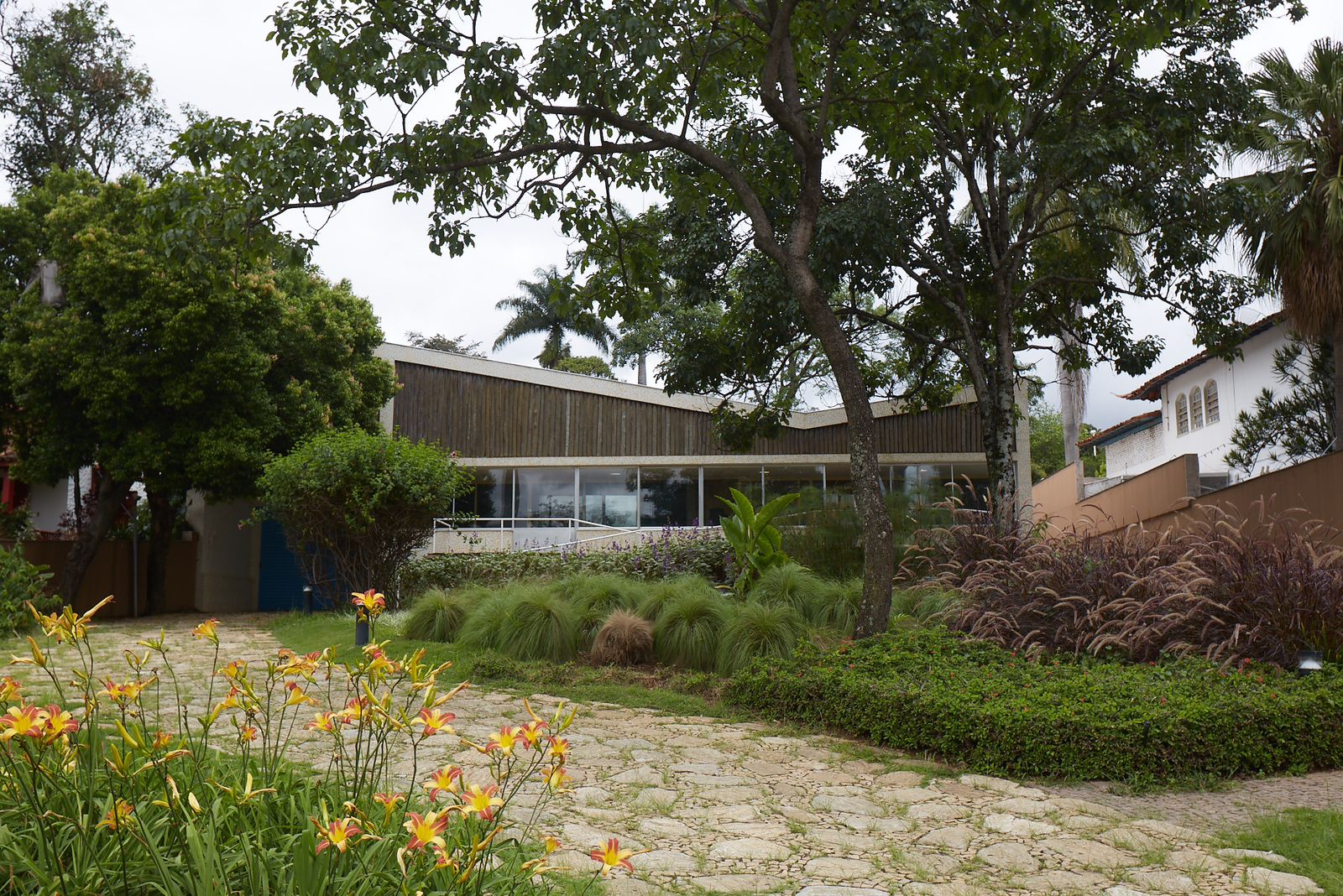 Imagem apresenta a fachada do Museu Casa Kubitschek. Uma entrada com chão de pedra, cercado pelo jardim. É possível notar árvores ao fundo e no centro da foto há a Casa Kubitschek, construção branca, com janelas em vidro. 