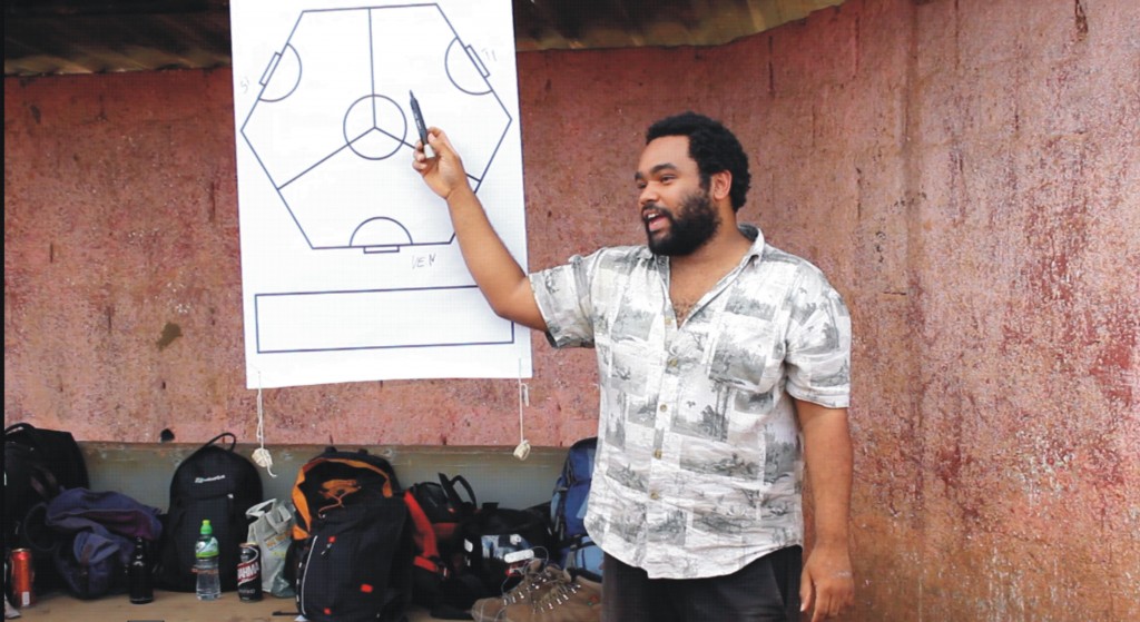 Um homem negro de barba, camisa branca está de pé. Ele aponta para um quadro branco com uma caneta na mão. No quadro branco há um desenho.