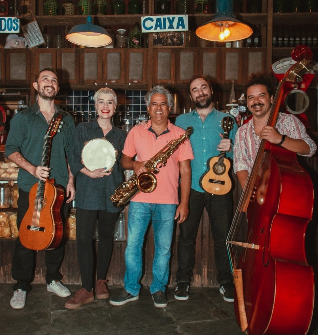 Grupo Choro do Jura posa para câmera enquanto seguram instrumentos. Ao todo são cinco pessoas, sendo quatro homens e uma mulher. O primeiro segura um violão, a segunda segura um pandeiro. O terceiro integrante da banda segura um saxofone, já o quarto musico tem um cavaquinho. Por fim, o último homem está com um violoncelo. 