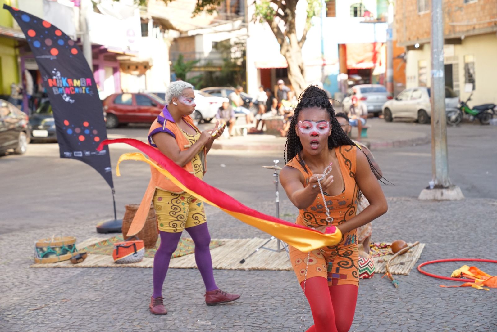 A imagem revela em primeiro plano uma mulher negra, com pintura facil. Ela está se apresentando segurando uma fita colorida, amarela e vermelha. A artista usa uma roupa laranja e cabelos de trança. Ao fundo, é possível notar outra mulher com roupas coloridas e pintura facial. Essa usa o cabelo raspado e loiro. 