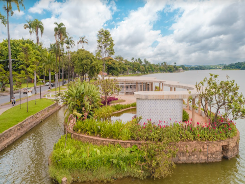 Fachada lateral da casa do baile, cercada pelo jardim florido e com árvores. Ao fundo está a Lagoa da Pampulha