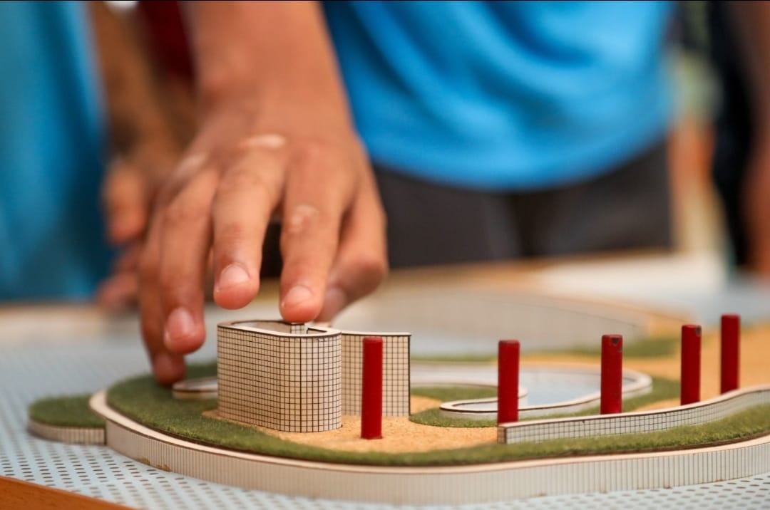 A mão de uma criança toca uma escultura, maquete de um prédio. A obra tem cores bege, vermelha e tons verdes. 