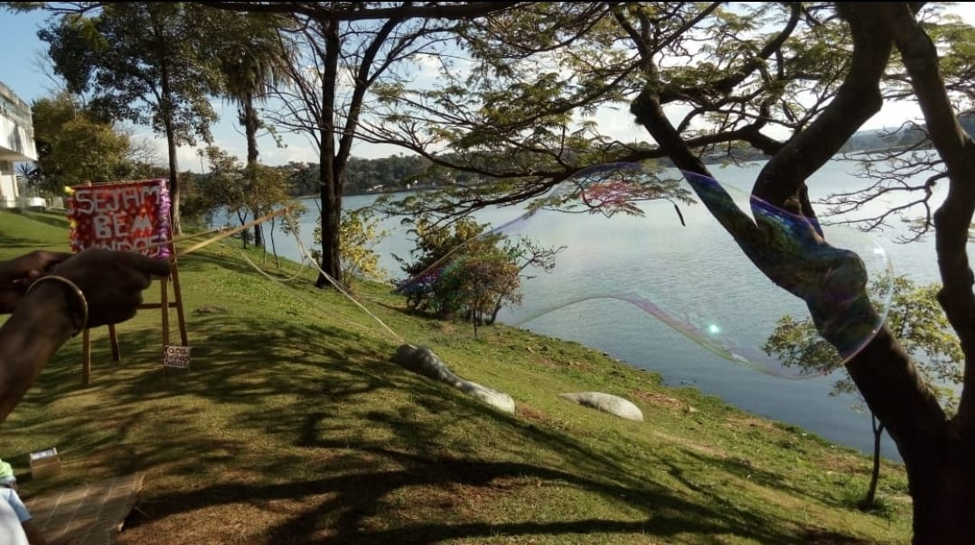 Mãos negras seguram dois palitos amarelos, usados para fazer uma grande bola de sabão. A imagem apresenta um gamado e árvores, em um dia ensolarado. Ao fundo, parte da Lagoa da Pampulha. 