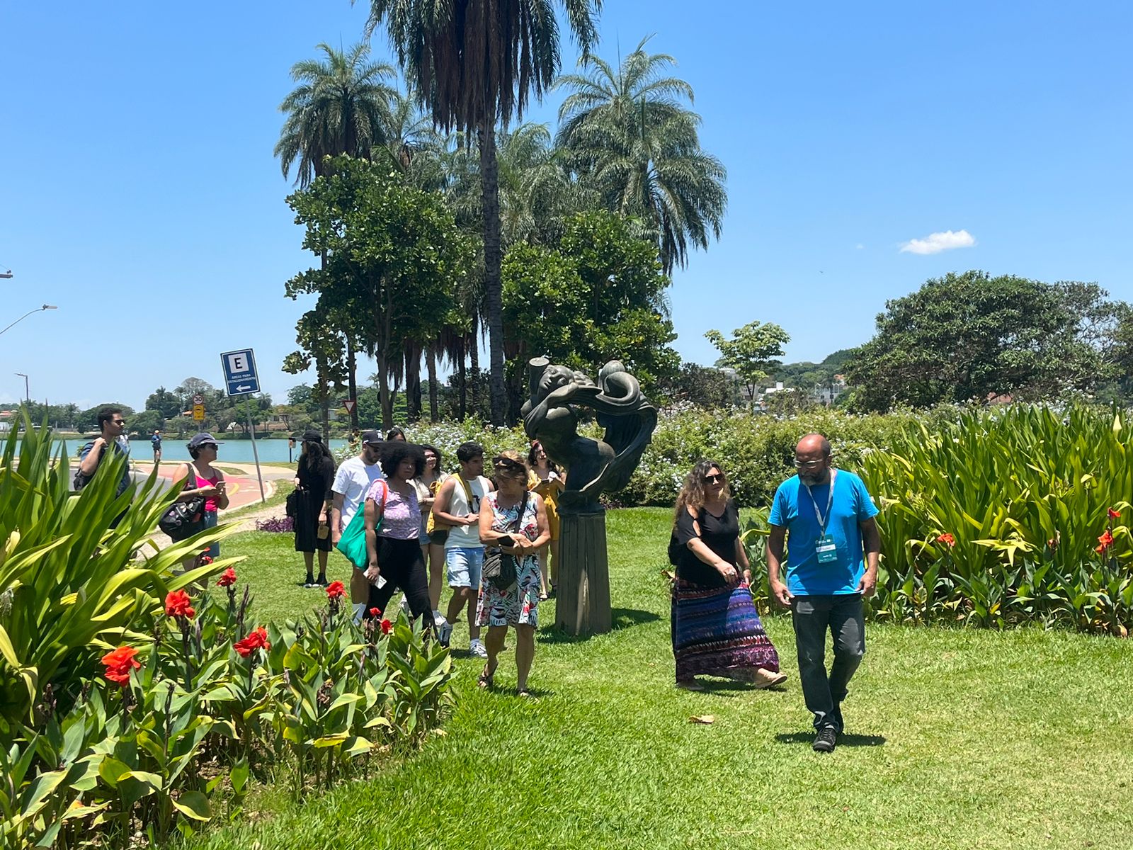 Grupo de pessoas caminha pelos jardins do Museu de Arte da Pampulha. O dia está ensolarado, a grama verde e o céu azul. Há coqueiros ao fundo