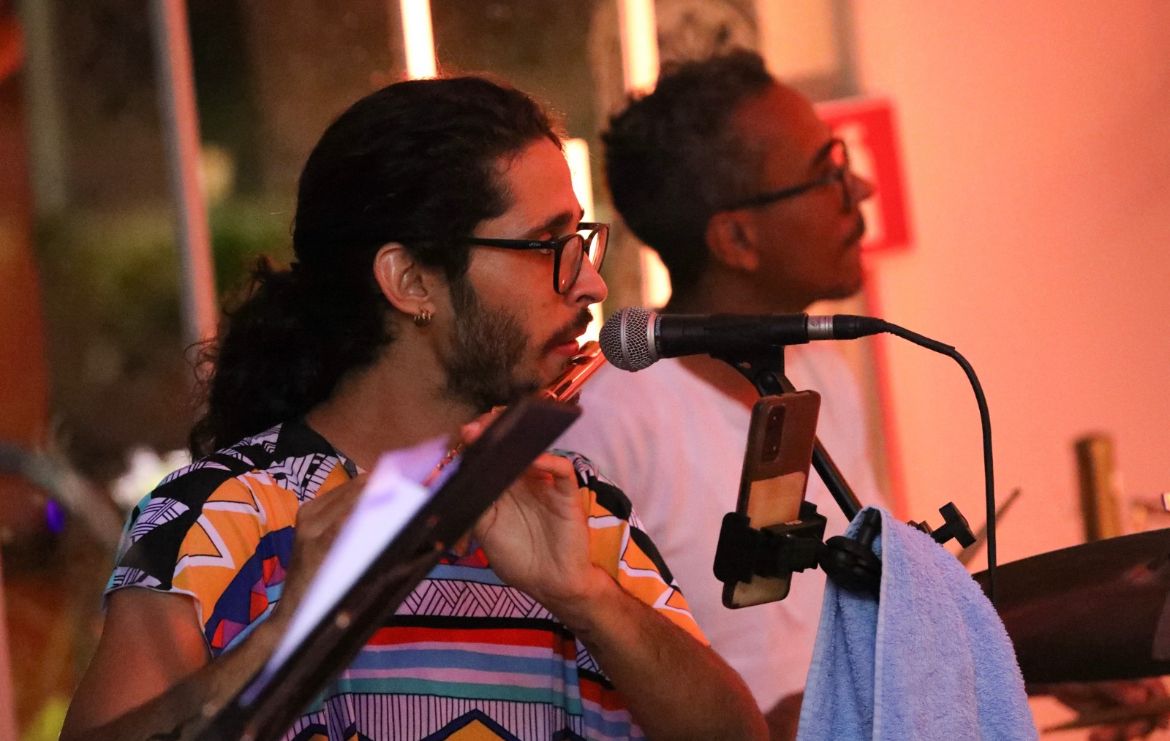 Em primeiro plano, um homem branco de óculs, cabelo comprido amarrado e baba, aparece tocando um instrumento de sopro. Ao fundo há um homem negro de óculos e camiseta branca