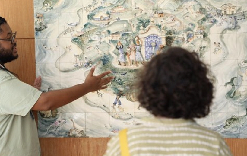 Uma mulher, que está de costas para câmera, observa um quadro feito de azulejos coloridos, enquanto um homem negro faz a explicação da obra. Ela possui cabelos curtos e está usando roupa verde e branca. Ele possui cabelos longos e trançados. 