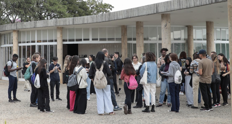 Um Grupo de pessoas aguardam em pé na porta da Casa do Baile. Elas usam mochilas. 