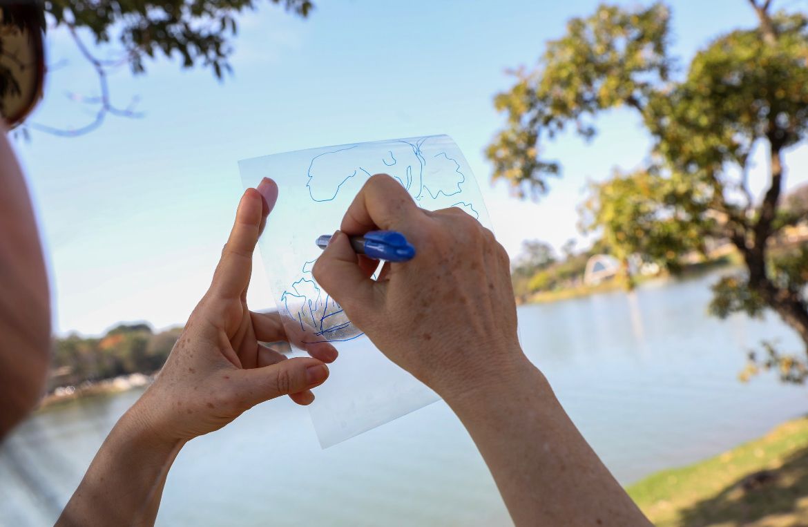 Enquanto uma mão segura uma folha transparente, a outra desenha com caneta azul. O papel está levantado para o alto, podendo ser observado a paisagem ao fundo: a lagoa da Pampulha, um pedaço gramado e arvores nas laterais da imagem 