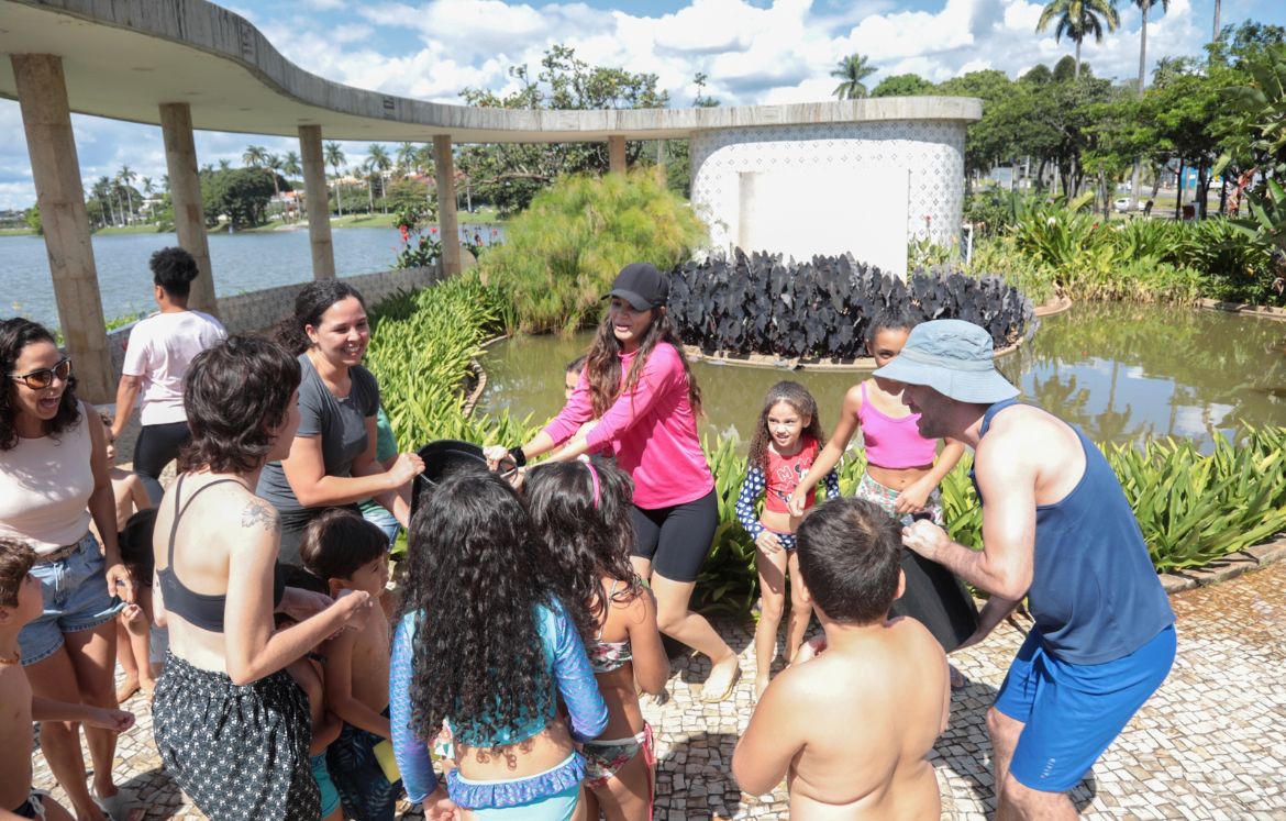 Grupo de crianças e alguns adultos, com trajes de banho, seguram baldes com água. Ao fundo é possível observar os jardins e estrutura da Casa do Baile