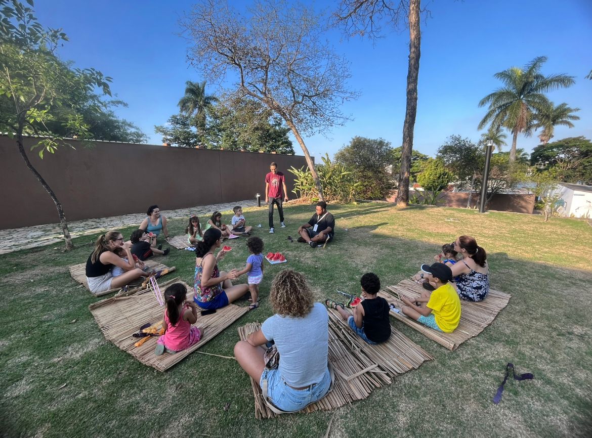 grupo de crianças e alguns adultos, todos sentados em esteiras de bambu, nos gramados do jardim MCK