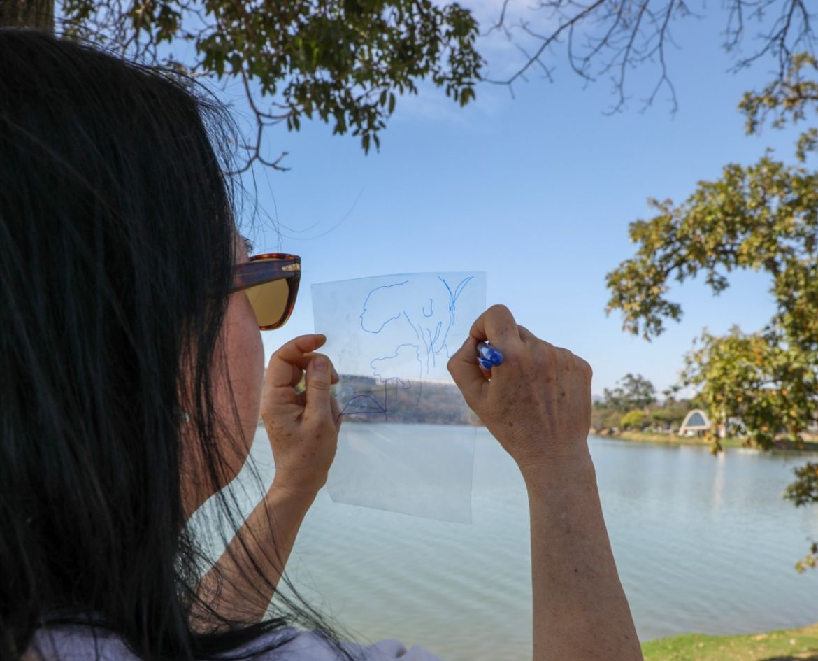 Uma mulher, de costas para câmera, segura um papel transparente e faz um desenho nele. Ela está posicionada de frente para a Lagoa da Pampulha 