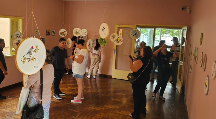 Diversas pessoas em pé em uma sala com paredes rosas observam os bordados pendurados em todo o teto. Cada bordado apresenta um passarinho diferente 