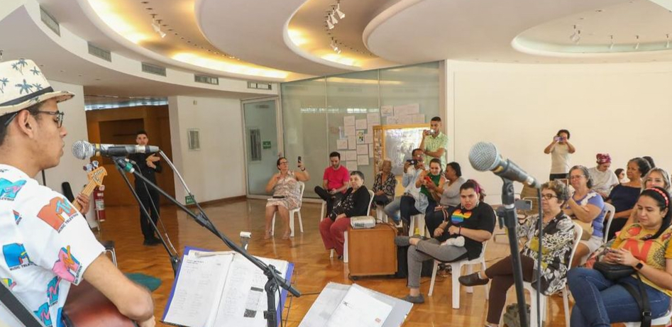 Um homem de blusa estampada e chapéu toca violão e canta para uma plateia sentada 