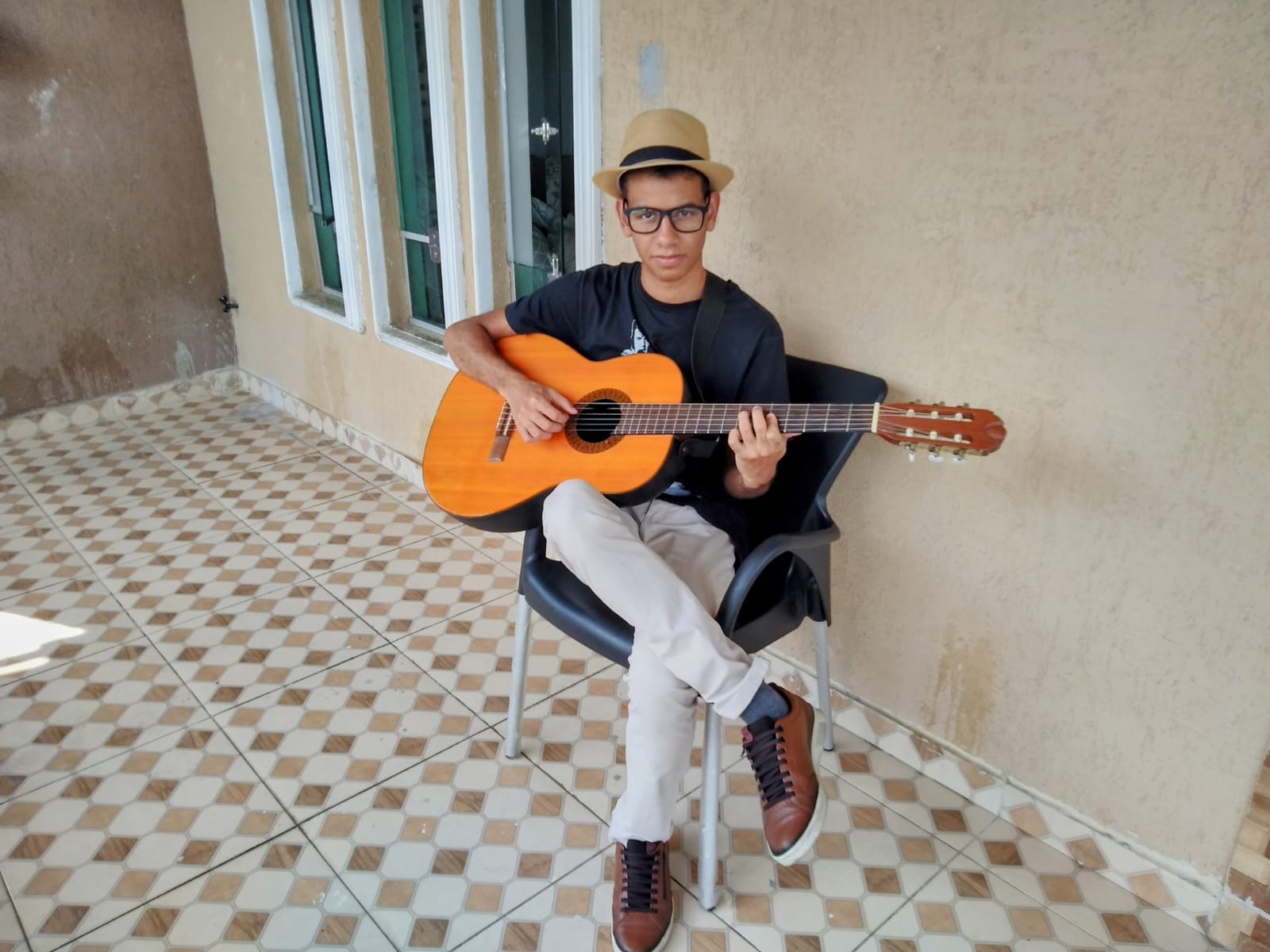 Um homem sentado tocando violão. Ele está olhando para câmera, usando uma calça clara, uma blusa preta e chapéu. 