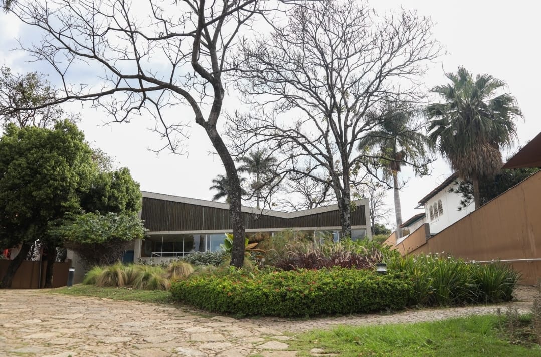 Imagem apresenta os jardins do Museu Casa Kubistschek. A foto mostra árvores, gramados e arbustos. Ao fundo, é possivel notar a fachada do Museu 