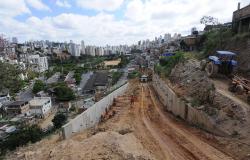 Homens trabalham na instalação de um grande muro de contenção no Aglomerado da Barragem Santa Lúcia. Mais acima vemos rua sendo feira com escavadora ao fundo, outro muro de contenção já pronto e um trator. Ao fundo, casas do Aglomerado e prédios de BH. 