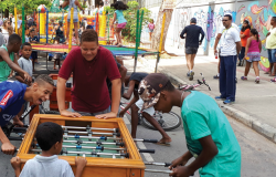 Quatro garotos jogam pebolim, ao fundo uma cama elástica e pessoas passando. 