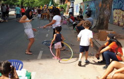 Três crianças brincam de bambolê, ao lado, um garoto passa; duas pessoas estão sentadas na calçada e ao fundo, mais gente transita. Foto diurna.