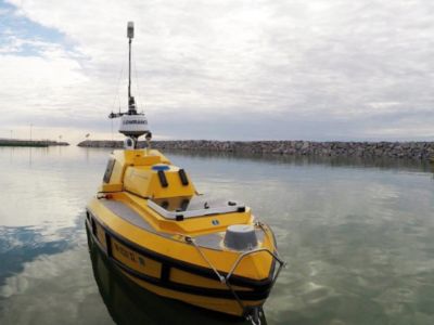 a small yellow vessel with a tall antennae floats on the surface of the water in a harbor with a rock breakwall